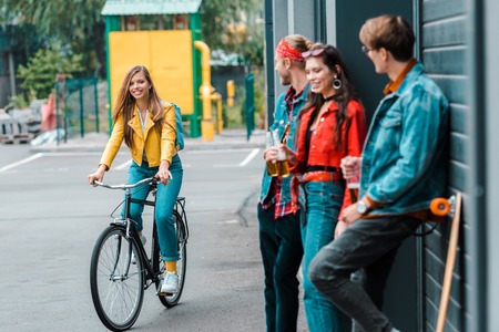 attractive stylish girl riding bike near friends with beer bottles on streetの写真素材