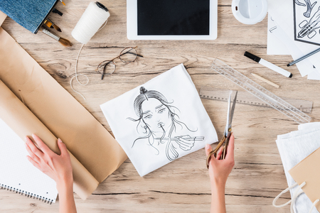 cropped image of female fashion designer with scissors sitting at table with paintings and digital tabletの写真素材