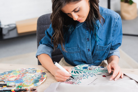 selective focus of female fashion designer painting on jacket at working table in clothing design...