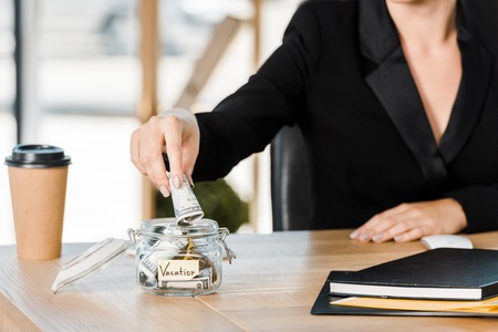 cropped image of businesswoman putting money in glass jar with note Vacation in office, travel conceptの写真素材