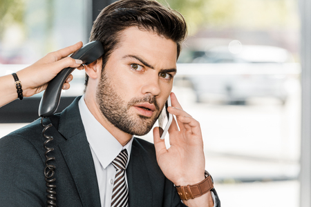 shocked businessman talking by smartphone and secretary giving him handset of stationary telephoneの写真素材