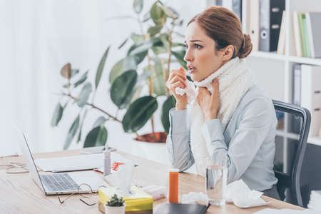 adult sick businesswoman with flu sitting at workplace in scarfの写真素材