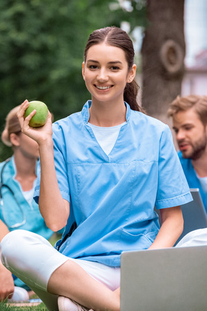 smiling medical student holding ripe green apple and sitting with laptopの写真素材