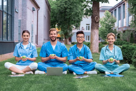 happy multicultural medical students sitting on grass and holding applesの写真素材