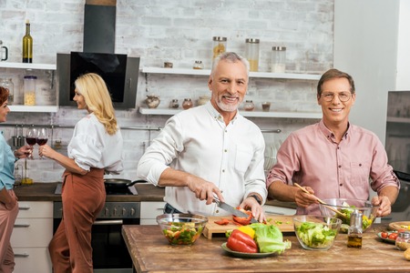 handsome smiling men preparing salad for dinner at home, women talking with wineの写真素材