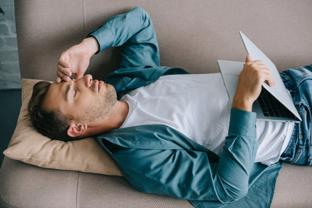 young man with headache holding laptop while lying on sofaの写真素材