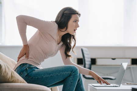 side view of young woman suffering from back pain while using laptop at homeの写真素材