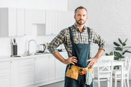 handsome plumber in overalls and tool belt looking at camera in kitchenの写真素材