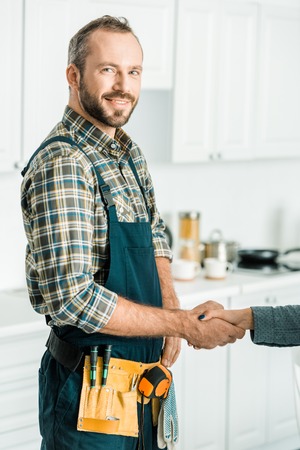 smiling handsome plumber and customer shaking hands in kitchenの写真素材