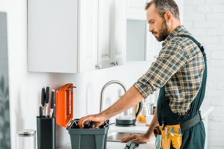 side view of handsome plumber taking tools from toolbox in kitchenの写真素材
