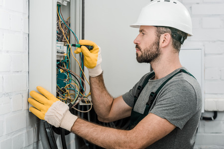 side view of handsome bearded electrician repairing electrical box and using screwdriver in corridorの写真素材
