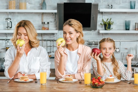 happy little child with mother and grandmother with pancakes and healthy apples for breakfast at...