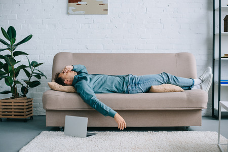 young man with headache lying on sofa, laptop on carpet at homeの写真素材