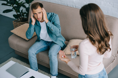young man with headache looking at woman holding glass of water and pillsの写真素材
