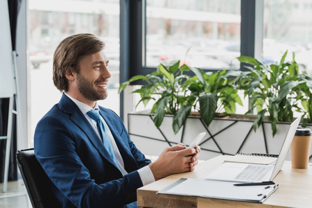 side view of smiling young businessman using smartphone and laptop in officeの写真素材