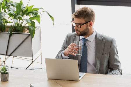 young businessman holding glass of water and looking away while using laptop in officeの写真素材