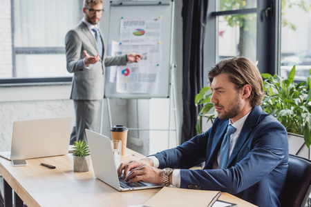 young businessman using laptop while colleague standing near whiteboard behindの写真素材