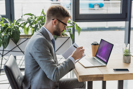 young businessman in suit and eyeglasses taking notes in notebook and using laptop with business graphsの写真素材