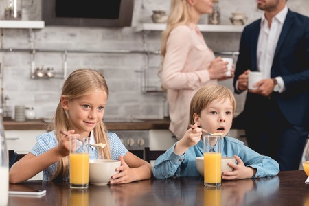 cute little kids having breakfast together at kitchen while parents drinking coffee on backgroundの写真素材