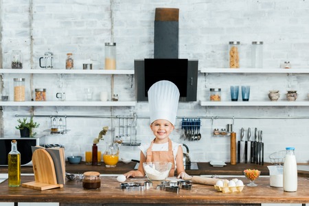 adorable kid in chef hat and apron cooking dough and smiling at cameraの写真素材