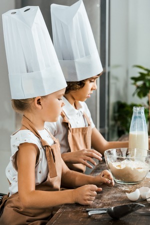 side view of adorable kids in chef hats and aprons preparing dough for cookies in kitchenの写真素材
