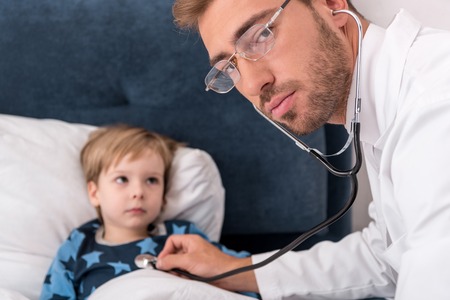 pediatrician litening childs breath with stethoscope and looking at camera while he lying in bedの写真素材