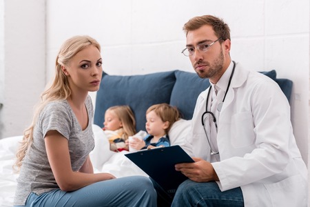 pediatrician and mother looking at camera while sick kids lying in bed on backgroundの写真素材