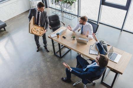 high angle view of young businessman holding pizza in box and looking at coworkers in officeの写真素材