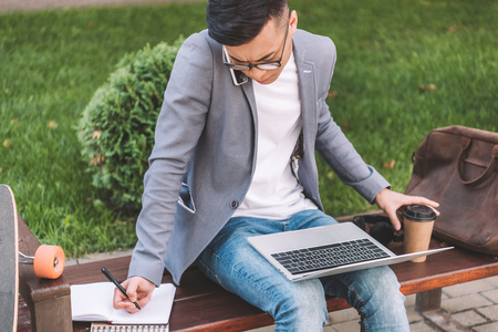 asian freelancer writing in planner while using smartphone and laptop on benchの写真素材