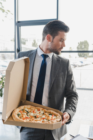 handsome young businessman in suit holding box with pizza in officeの写真素材
