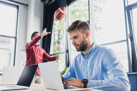 focused young businessman using laptop while colleague playing with rugby ball behind in officeの写真素材