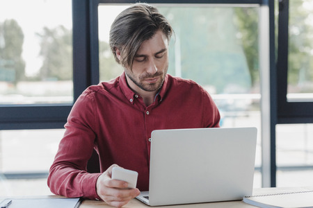 businessman holding smartphone and using laptop while working in office