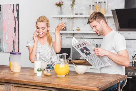 girlfriend talking by smartphone and gesturing during breakfast in kitchen, boyfriend reading newspaperの写真素材