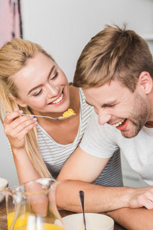 smiling girlfriend feeding laughing boyfriend with cornflakes in kitchenの写真素材