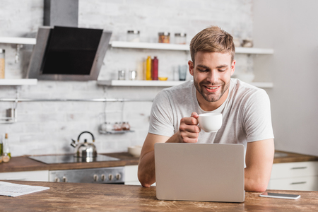 handsome smiling man holding cup of coffee and looking at laptop in kitchenの写真素材