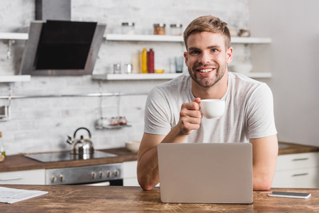 handsome smiling man holding cup of coffee and looking at camera in kitchenの写真素材