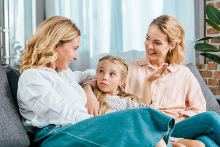 happy child with mother and grandmother sitting on couch at homeの写真素材