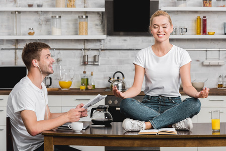 smiling young man reading newspaper and looking at happy girlfriend meditating on kitchen table