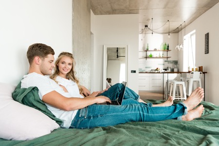 smiling young couple with digital laptop resting on bed at homeの写真素材