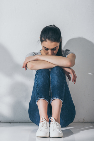 full length view of sad stressed girl sitting with hands on knees on floorの写真素材