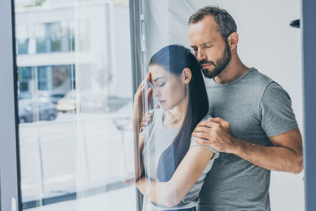 bearded man supporting sad stressed woman with closed eyes standing near windowの写真素材