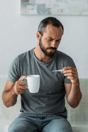 sick bearded middle aged man holding cup and looking at thermometerの写真素材