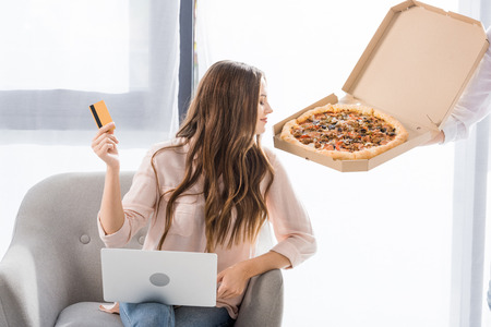 partial view of man with pizza in paper box and young woman with credit card and laptop at home, shopping online conceptの写真素材