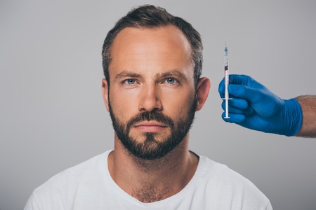doctor holding syringe in hand and man with alopecia looking at camera isolated on greyの写真素材