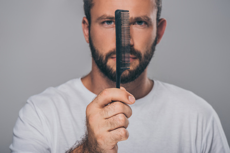 bearded man holding comb and looking at camera isolated on grey