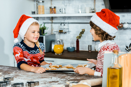 adorable happy children in santa hats holding baking tray with ginger cookies and smiling each otherの写真素材