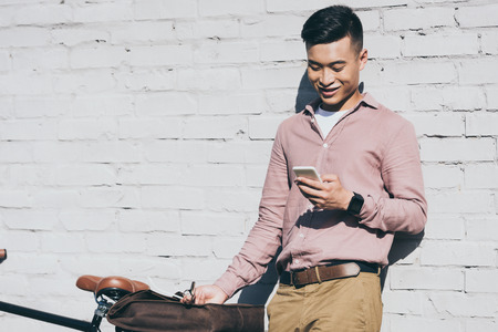 smiling asian man using smartphone while standing near bicycle on streetの写真素材