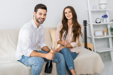 happy young couple celebrating with bottle of champagne and glasses on sofa at homeの写真素材