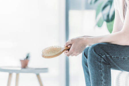 cropped shot of young woman holding hairbrush while sitting indoors, hair loss conceptの写真素材