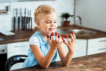 cute happy kid sitting at table and looking at ripe fresh raspberries on fingersの写真素材